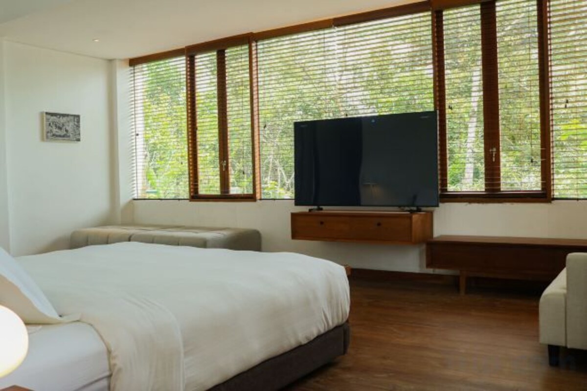 A bedroom with a bed, a television on a stand, a bench, and large windows with blinds.