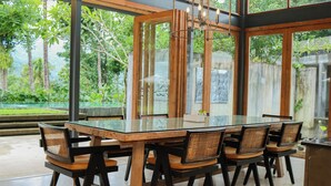 Uma área de refeições moderna, com mesa de vidro e cadeiras pretas, cercada por grandes janelas que oferecem vista para vegetação e uma parede de pedra.