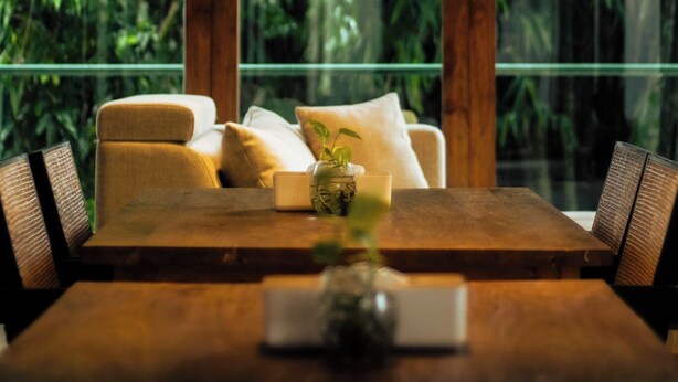 Uma mesa de jantar de madeira com cadeiras, um sofá e um guarda-corpo de vidro com vista para um jardim verdejante.