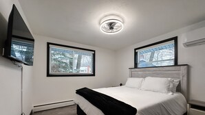 A bedroom with a bed, a nightstand, a wall-mounted TV, and two windows with blinds.