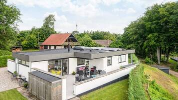 Una casa moderna de dos pisos con techo plano, ventanas amplias y un patio con mobiliario exterior.