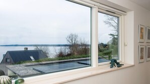 A modern room with a large window overlooking a body of water, a small shelf with decorative items, and a wall with framed pictures. - 5 Star Holiday Home in Borkop (Borkop)