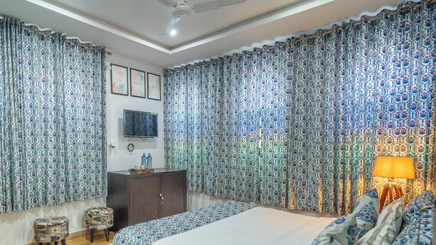 A bedroom with a large bed, curtains, a ceiling fan, a TV, and a bedside table. - Footprint Hostel Udaipur (Udaipur)