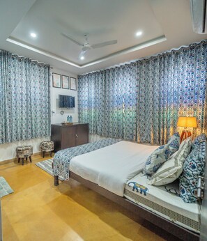 A bedroom with a large bed, curtains, a ceiling fan, a TV, and a bedside table. - Footprint Hostel Udaipur (Udaipur)