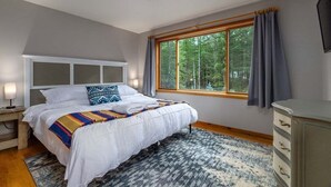 Une chambre à coucher comprenant un lit, une table de chevet, une commode, une fenêtre avec des rideaux et une vue sur les arbres.