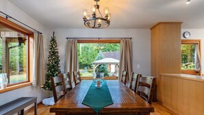 Un coin repas avec une table en bois, des chaises, un lustre, un sapin de Noël et une vue sur l’extérieur.