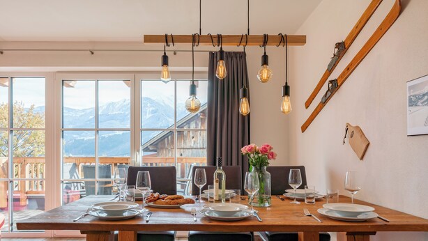 Ein Essbereich mit Holztisch, gedeckt für vier Personen, große Fenster mit Blick auf die Berge und einem Skiständer an der Wand.