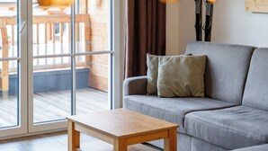 A living room with a grey sofa, a wooden coffee table, and a glass door leading to an outdoor area with wooden furniture.