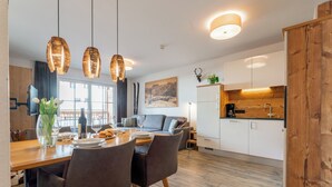 A modern kitchen with wooden cabinets, a dining area with a wooden table and chairs, and a living area with a sofa and a painting on the wall.