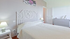 Une chambre à coucher avec un lit blanc, une table de chevet avec une lampe, une commode et une porte donnant sur une autre pièce.