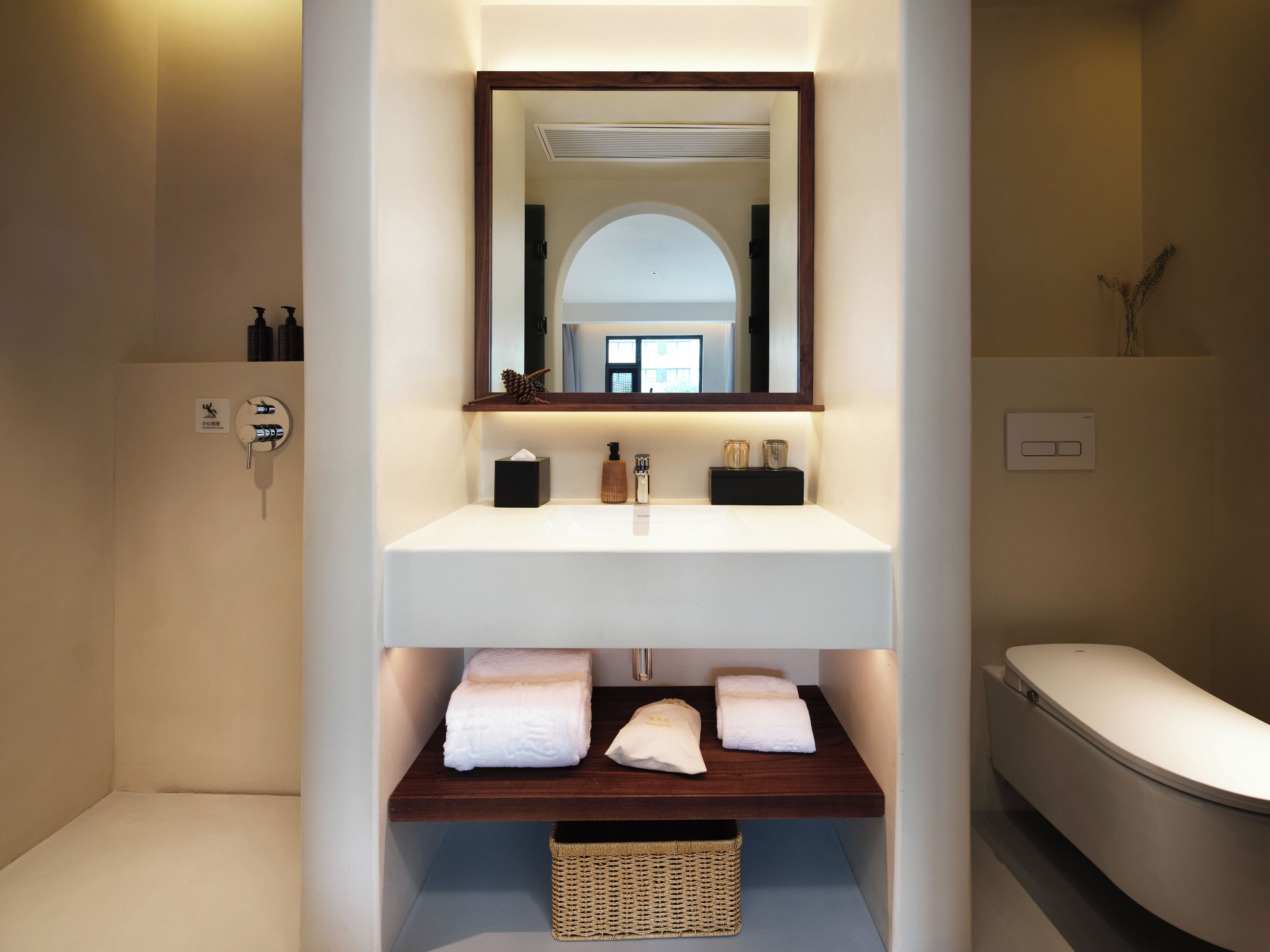 A modern bathroom with a white sink, a mirror above it, and a bathtub to the right. There are towels on the sink and a basket below it.