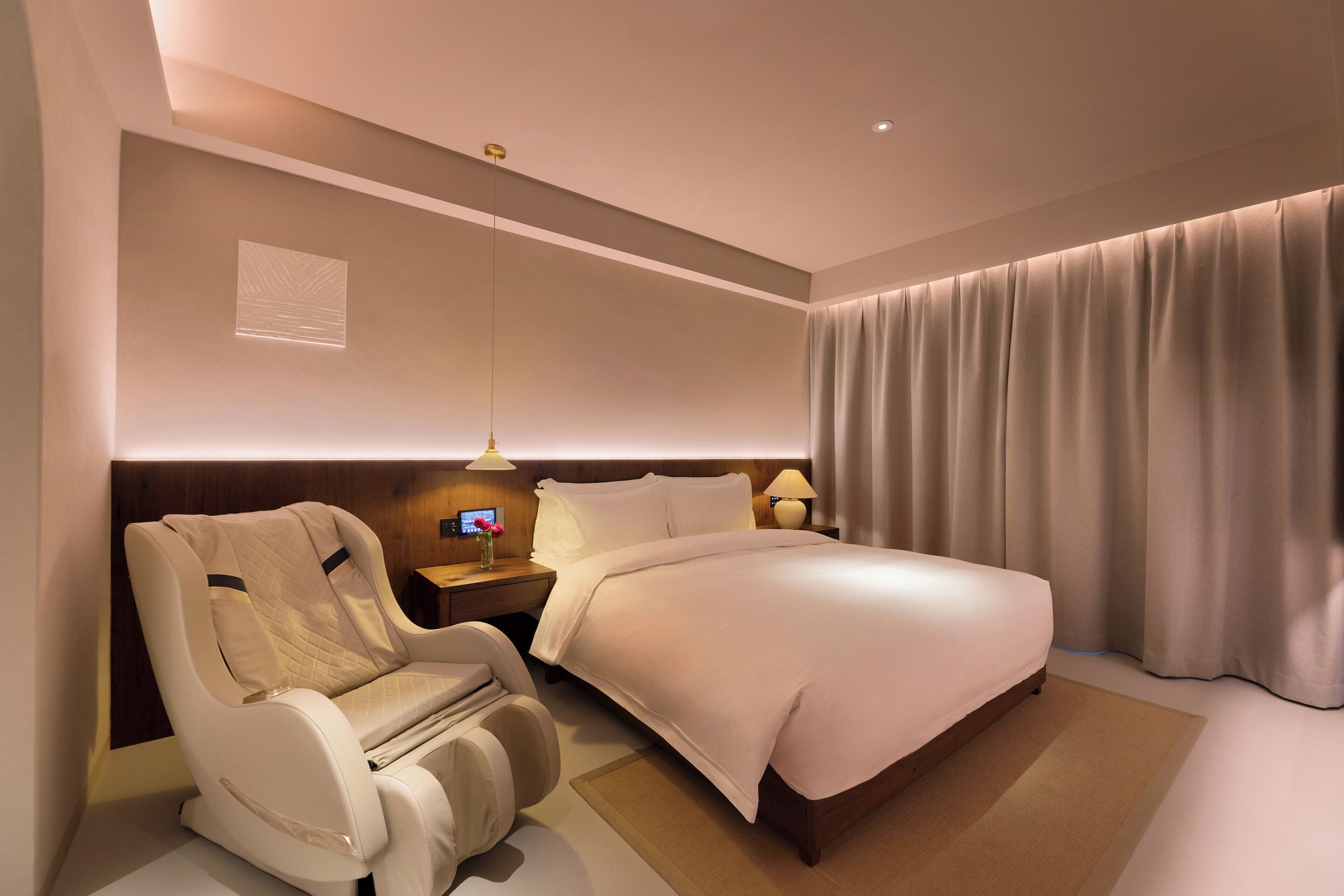 A modern hotel room with a large bed, a massage chair, a bedside table with a lamp, and a large window with curtains.