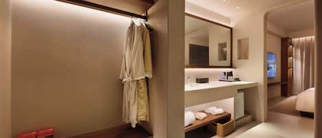 A modern hotel bathroom with a wooden vanity, a mirror, and a bed visible through an archway.