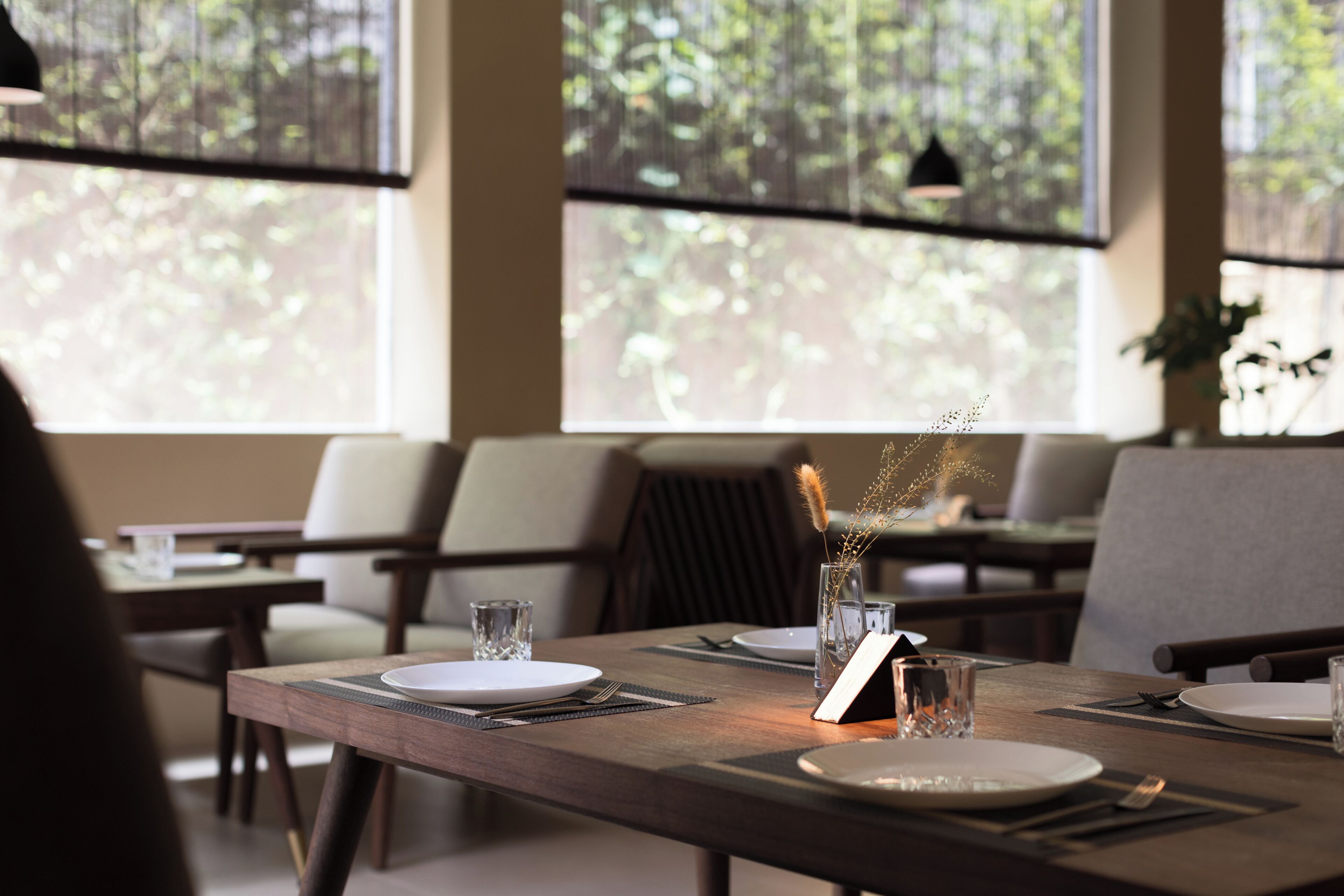 A dining area with tables set for two, featuring a modern design with wooden tables and chairs, and a view of greenery through large windows.