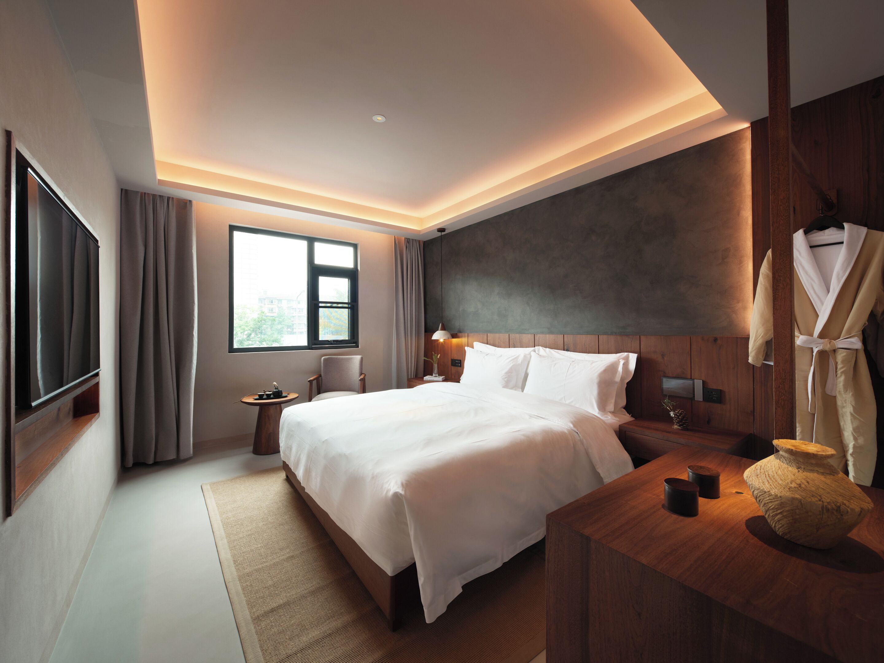 Een moderne hotelkamer met een groot bed, een houten hoofdbord, een nachtkastje met een vaas en een uitzicht op de stad door het raam.