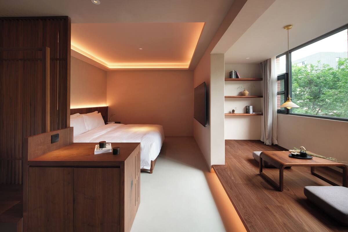 A modern hotel room with a large bed, a wooden desk, a TV, and a window with a view of greenery.