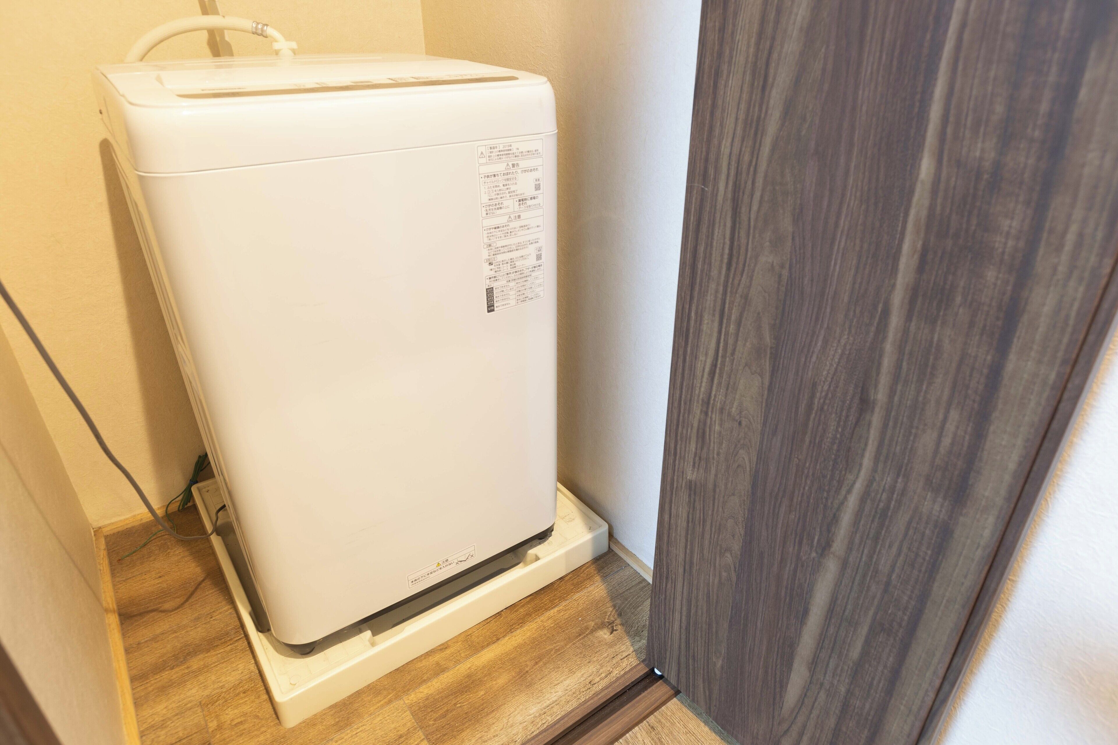 Un appareil électroménager compact et blanc, vraisemblablement un réfrigérateur, placé dans un coin d’une pièce dont le sol et la porte sont en bois.
