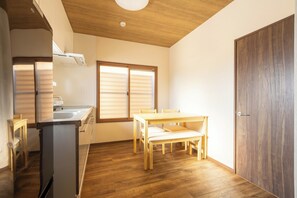 Une cuisine de taille réduite, avec une table et des chaises en bois, un réfrigérateur et une fenêtre équipée de stores.