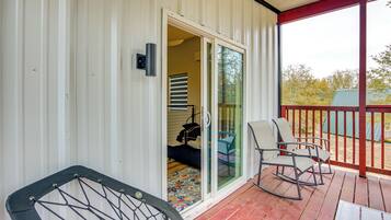 A covered porch with a red railing, a sliding glass door leading to a room with a patterned rug, and two rocking chairs.
