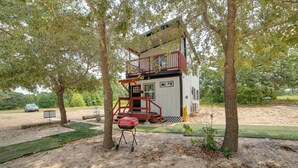A small, two-story cabin with a balcony, surrounded by trees and a barbecue grill. - Hike & Explore: 60-Acre Outdoor Retreat in Tyler! (Tyler)