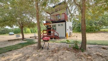A small, two-story cabin with a balcony, surrounded by trees and a barbecue grill.