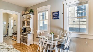 A dining area with a table, chairs, and a window with blinds. There is a sign on the wall and a shelf with decorative items.