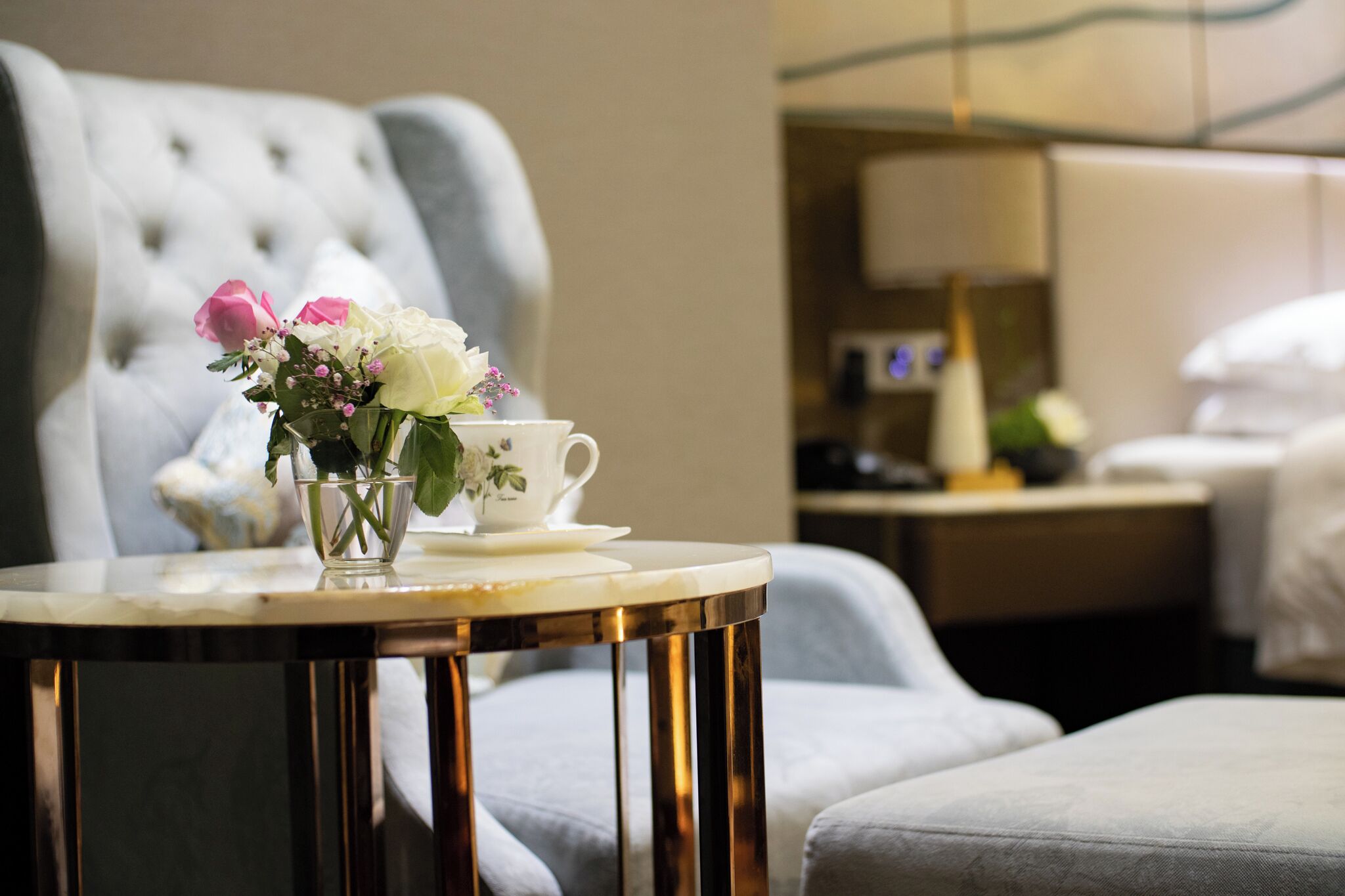 Ein Hotelzimmer mit einem geblümten Ohrensessel, einem kleinen runden Tisch mit einer weißen Tasse und einer Blumenvase, sowie einem Bett mit weißer Bettwäsche.