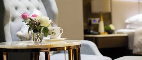 A hotel room with a tufted armchair, a small round table with a white cup and a vase of flowers, and a bed with white linens.