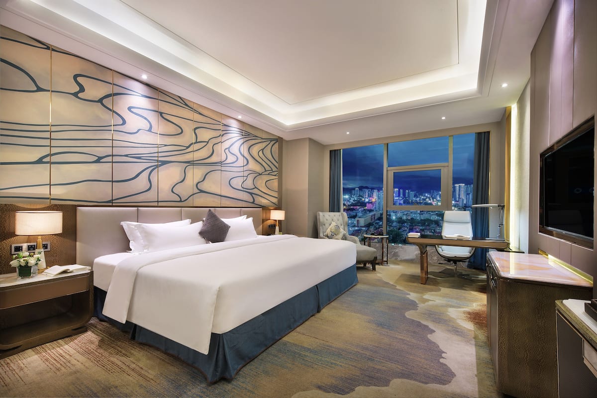 Habitación de hotel con una cama grande, un escritorio, una silla, un televisor y vistas a un paisaje urbano nocturno.