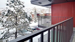 Ein Balkon mit Metallgeländer, der auf eine verschneite Landschaft, ein rotes Gebäude und geparkte Autos blickt.