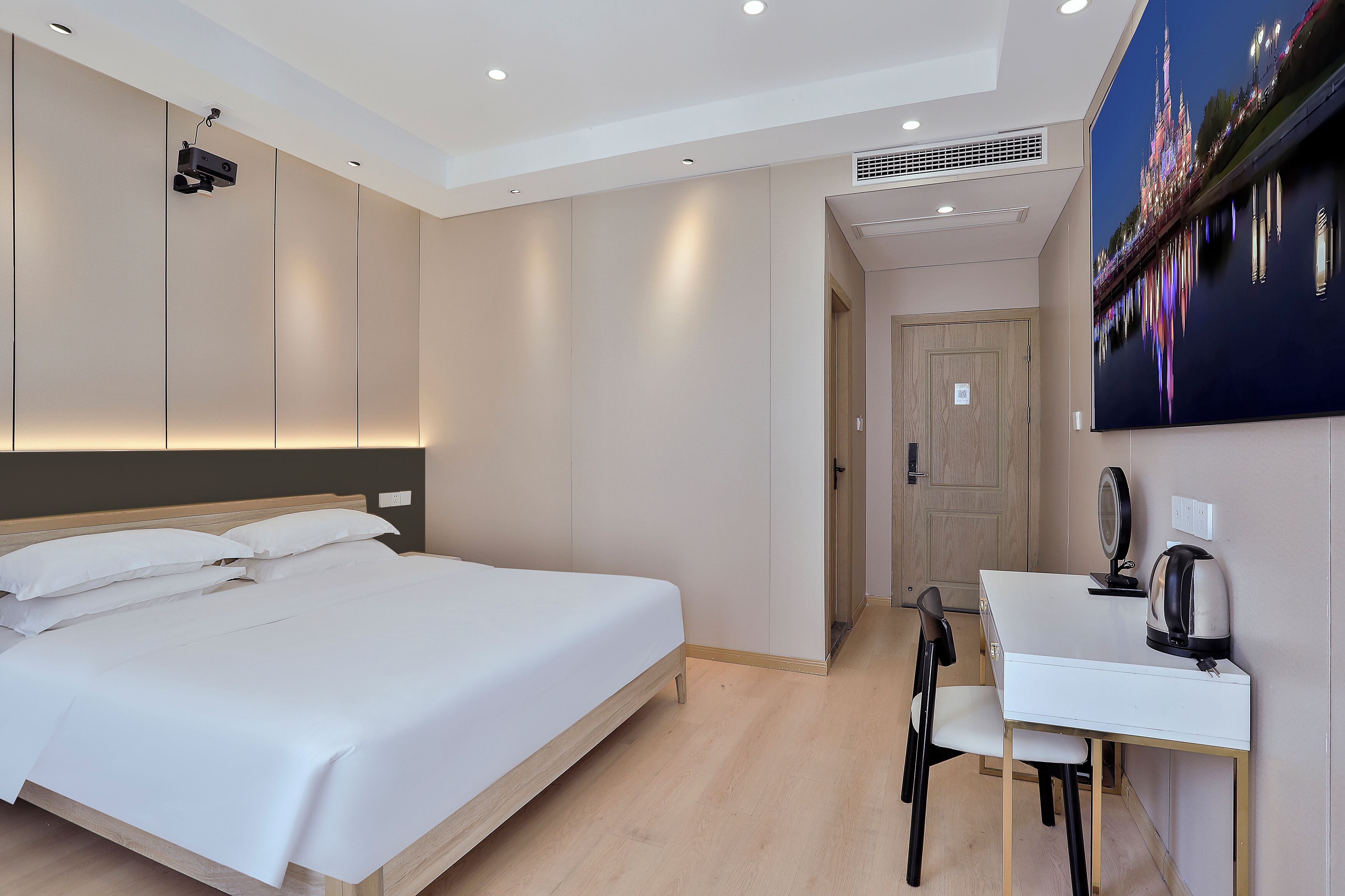 A modern hotel room with a large bed, a desk with a kettle, and a wall-mounted projector.