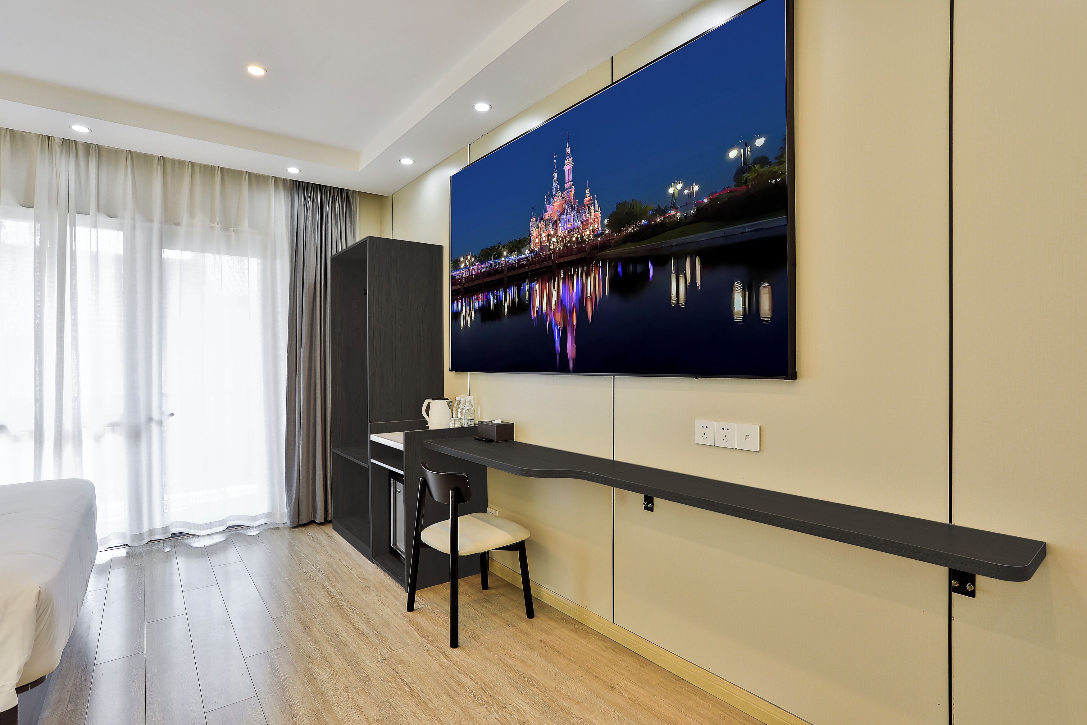 Ein modernes Hotelzimmer mit großem Bildschirm, der eine nächtliche Stadtansicht zeigt. Es gibt einen Schreibtisch mit Stuhl, ein Bett und ein großes Fenster mit durchscheinenden Vorhängen.
