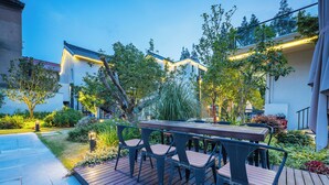 Une terrasse avec une table et des chaises en bois, entourée de verdure et illuminée par des guirlandes lumineuses.