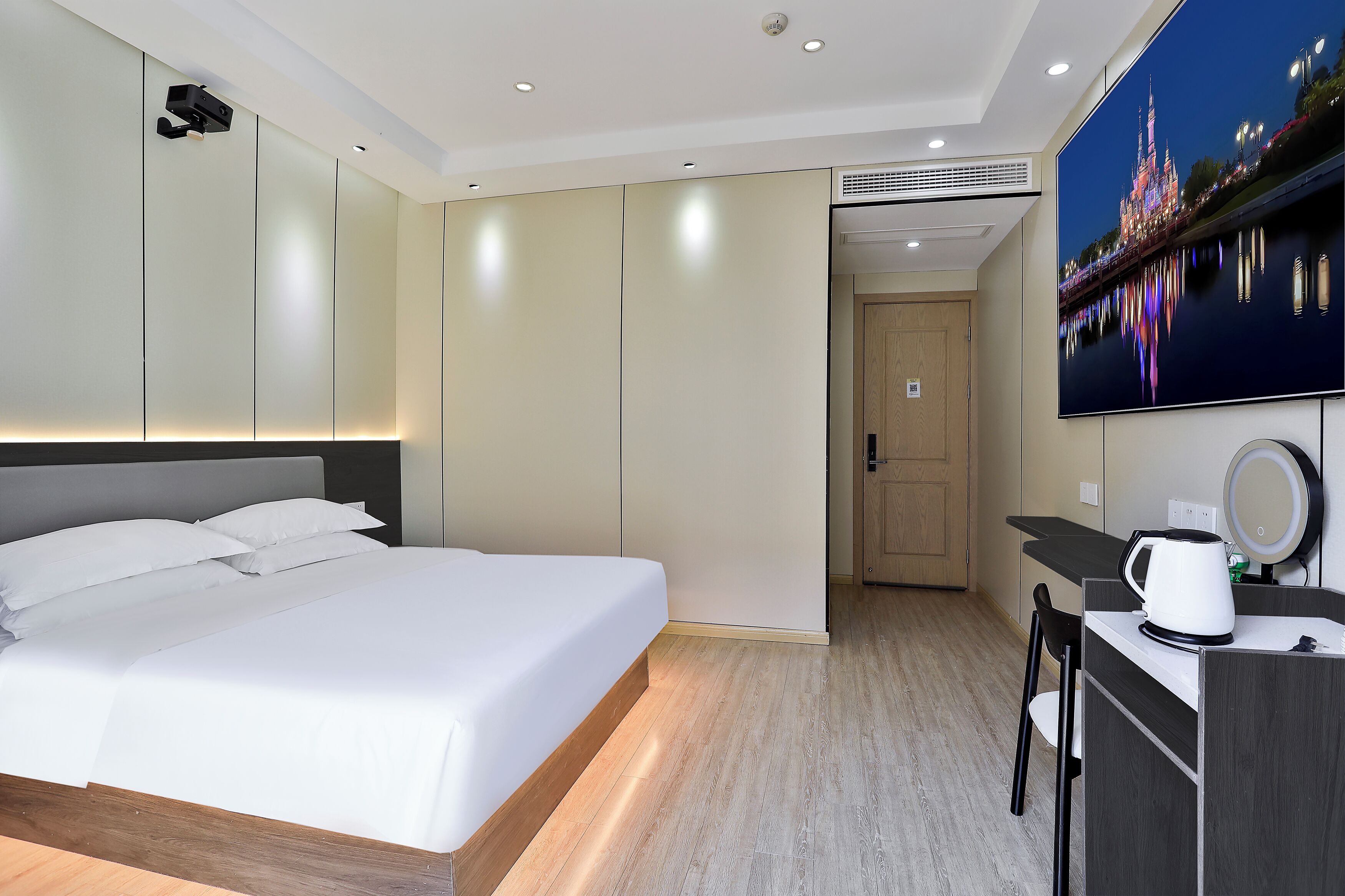 Una habitación de hotel moderna con una cama grande, un escritorio con cafetera, un televisor mostrando un paisaje urbano nocturno y una puerta que da a otra habitación.