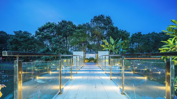 Une passerelle couverte de verre, dotée de rampes en métal, qui mène à l’entrée d’un bâtiment entouré d’arbres.