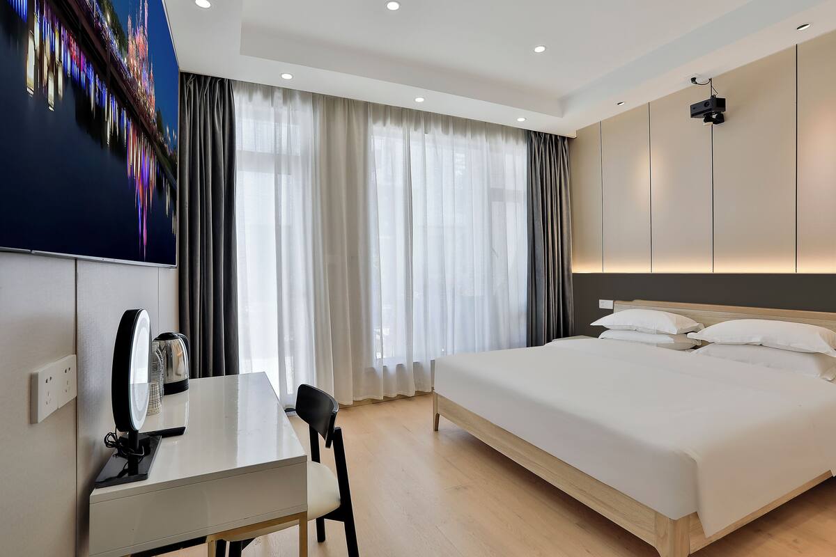 Une chambre d’hôtel moderne dotée d’un grand lit, d’un bureau avec un ordinateur, d’un téléviseur fixé au mur et d’une grande fenêtre avec des rideaux légers.