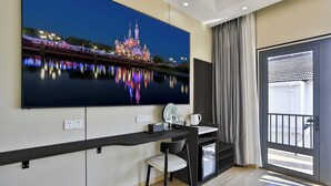 Une chambre d’hôtel dotée d’un grand téléviseur à écran plat fixé au mur, d’un bureau avec une chaise, d’une petite table avec une bouilloire et des tasses, d’une fenêtre avec des rideaux et d’une porte avec un portail en métal.