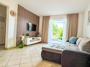 Ein Wohnzimmer mit einer Couch, einem an der Wand montierten Fernseher, einem Fenster mit Vorhängen und einem Fliesenboden.