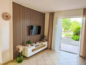 A modern living room with a TV mounted on a wooden panel wall, a white console table, and a green vase with decorative plants. - Apartment Mirta by Interhome (Krk/Šilo)