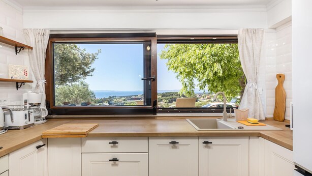 Een keuken met een groot raam, witte kastjes, een houten aanrechtblad en uitzicht op zee.