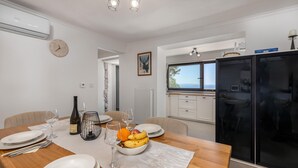 Een eetgedeelte met een houten tafel gedekt voor vier personen, witte borden, wijnglazen en een schaal met fruit. Een zwarte koelkast en een raam met uitzicht op zee zijn zichtbaar.