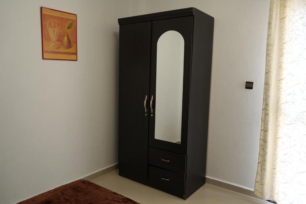 Une armoire de couleur sombre, dotée d’une porte miroir et de deux tiroirs, placée dans une pièce où l’on trouve un rideau à motifs et une œuvre d’art encadrée accrochée au mur.