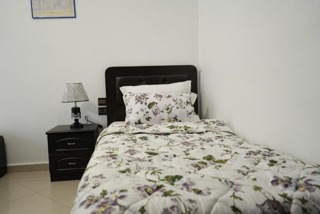 Un lit bien fait, recouvert d’une couette à motifs floraux et agrémenté d’un oreiller blanc, une table de chevet noire avec une lampe, et un cadre photo fixé au mur.