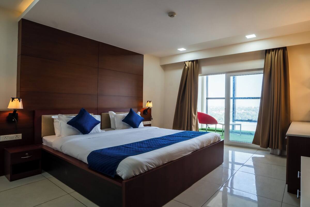 A modern hotel room with a large bed, bedside lamps, a desk with a mirror, and a view of the city through large windows.