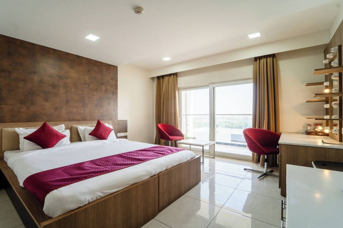 A modern hotel room with a large bed, two red chairs, a desk, and a balcony with a view.
