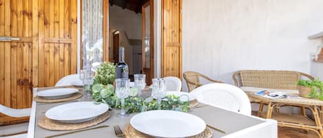 An outdoor dining area with a table set for two, surrounded by wicker chairs, and a wicker sofa with a magazine.