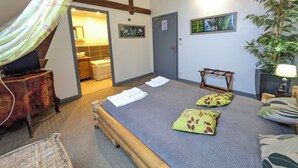 Un dormitorio con cama, una silla de madera, un escritorio con lámpara, una planta y un baño a la vista a través de una puerta abierta.