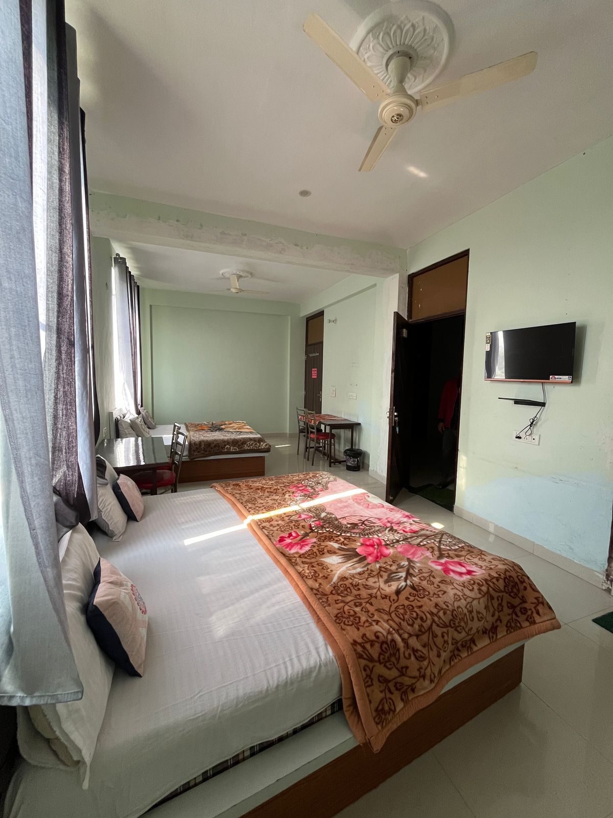 A hotel room with a large bed, a ceiling fan, a television mounted on the wall, and a dining area in the background.