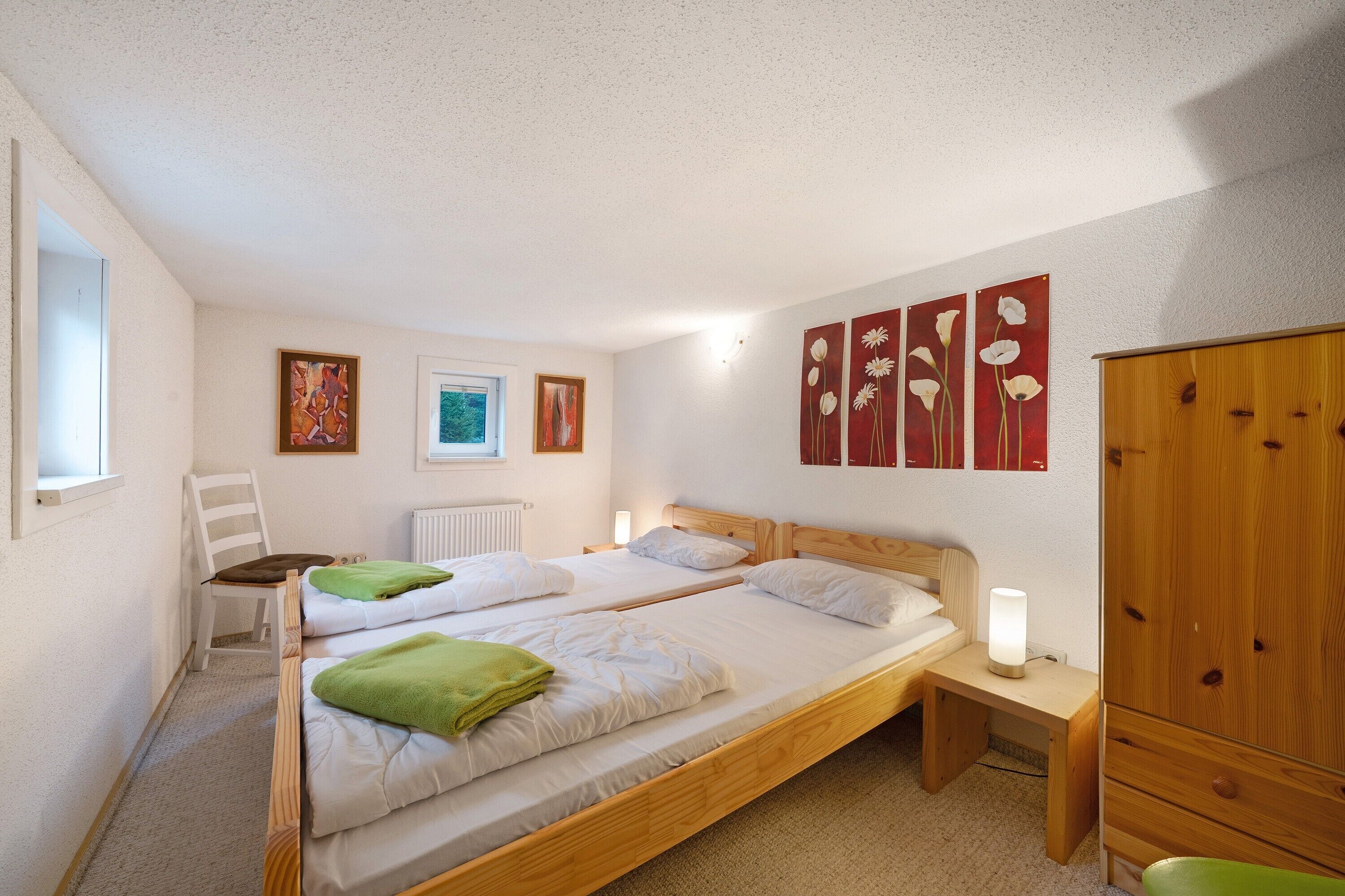 A bedroom with two single beds, a wooden wardrobe, a bedside table with a lamp, a chair, and artwork on the walls.