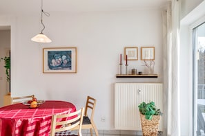 A dining area with a red checkered tablecloth, wooden chairs, a fruit bowl, a framed painting, a radiator, and a potted plant.